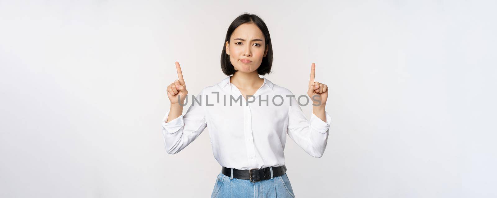 Image of skeptical asian lady grimacing, frowning upset, pointing fingers up, showing advertisement, standing over white background.