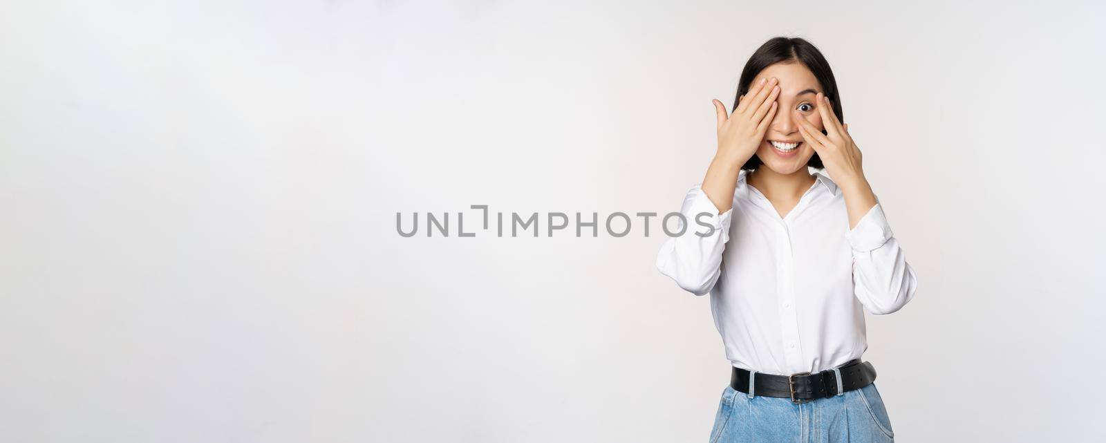 Image of young asian woman close her eyes and smiles, waits for surprise, anticipates, peeking through fingers, stands over white background.