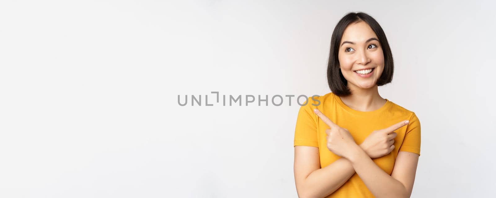 Cute asian girl pointing fingers sideways, showing left and right promo, two choices, variants of products, standing in yellow tshirt over white background.