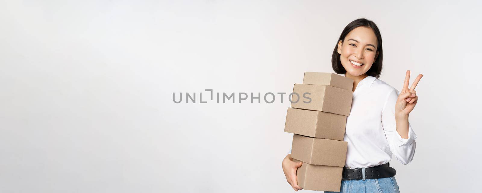 Smiling korean woman with boxes showing v-sign, peace gesture, standing over white background.