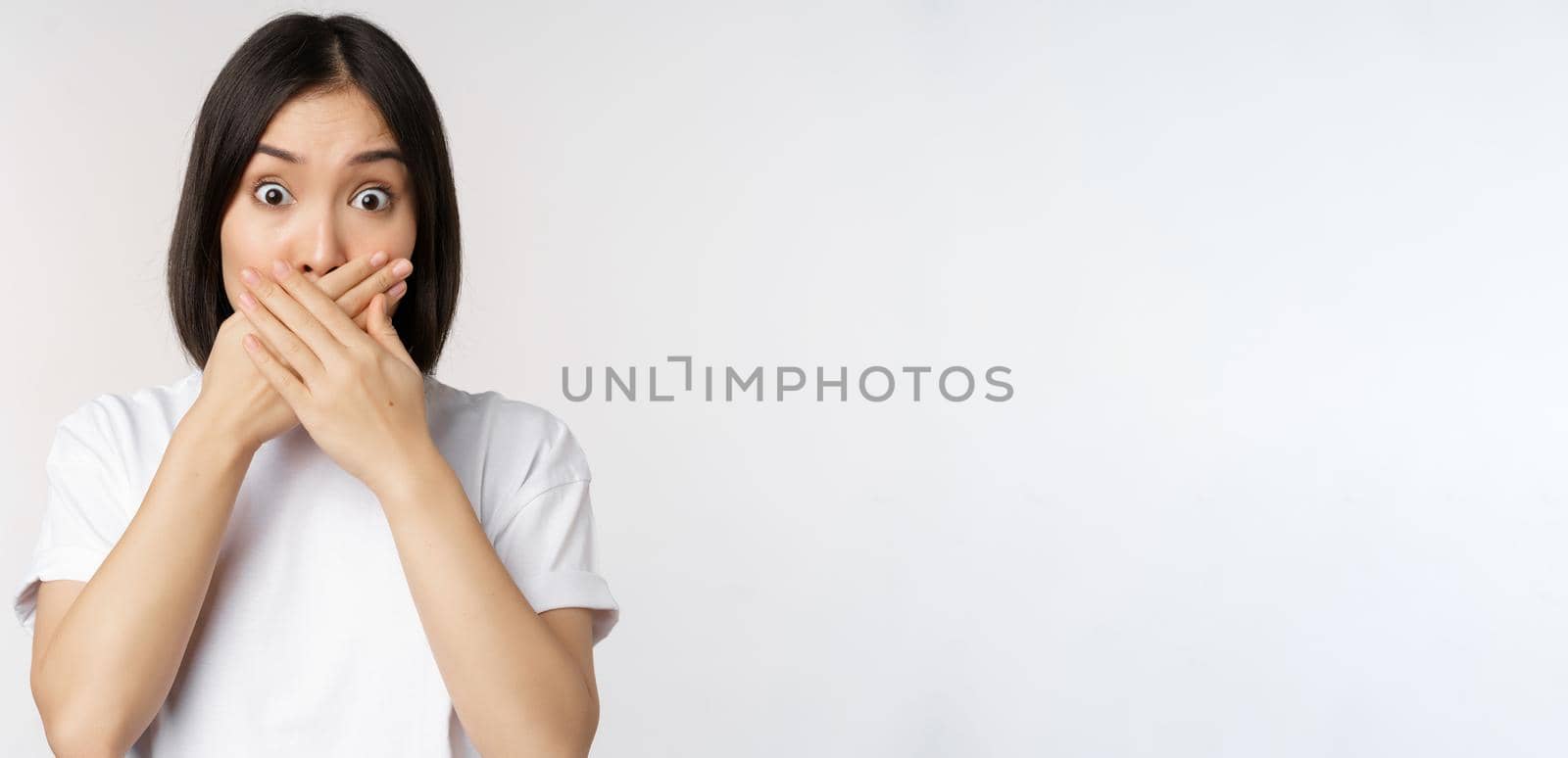 Image of shocked young asian woman shut mouth, looking startled, speechless, standing over white background by Benzoix