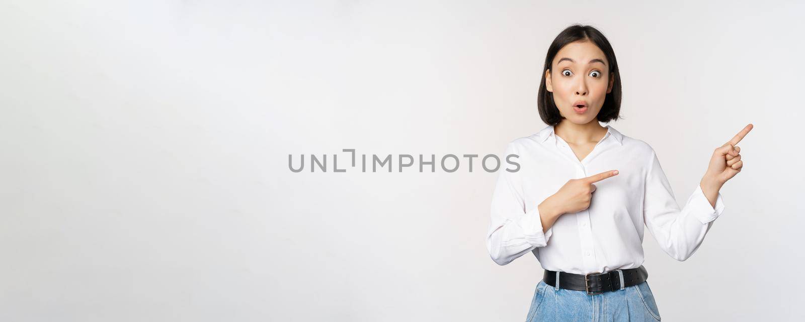 Surprised young woman pointing fingers right. Asian girl showing banner and looking enthusiastic, interested in advertising, standing over white background by Benzoix