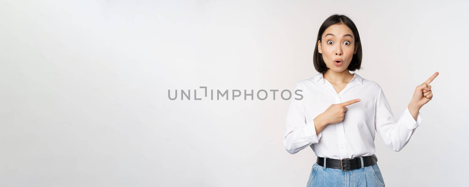 Surprised young woman pointing fingers right. Asian girl showing banner and looking enthusiastic, interested in advertising, standing over white background.