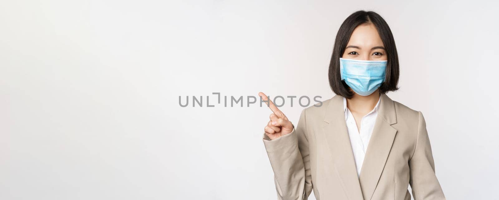 Coronavirus and work concept. Portrait of woman in medical face mask, pointing finger left, showing logo or banner, advertisement, white background.