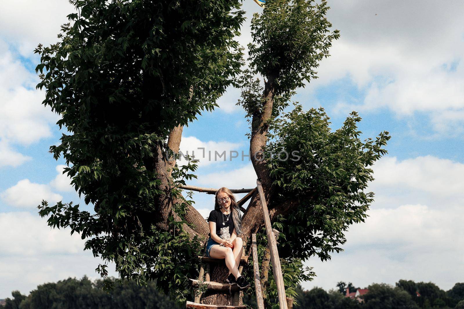 A young woman with a three-month-old Scottish Straight kitten sits on a beautiful green tree above the lake in summer. Walk, rest with a pet. The blonde is wearing short denim shorts, a black cotton T-shirt and glasses.