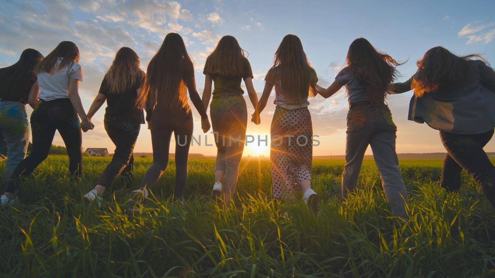 A group of girls walk towards the sun at sunset holding hands. by DovidPro