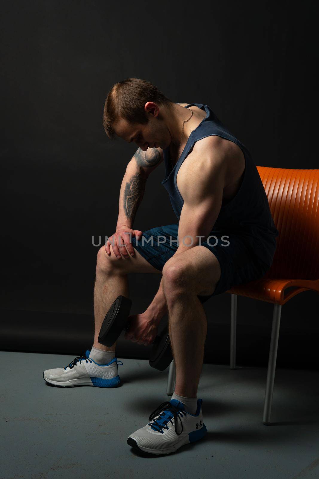 Man on black background keeps sits on a chair rocking his biceps dumbbells pumped up in fitness bodybuilding sexy sport, athlete weight strong athletic hand, person lifestyle. Young skin power, human fit