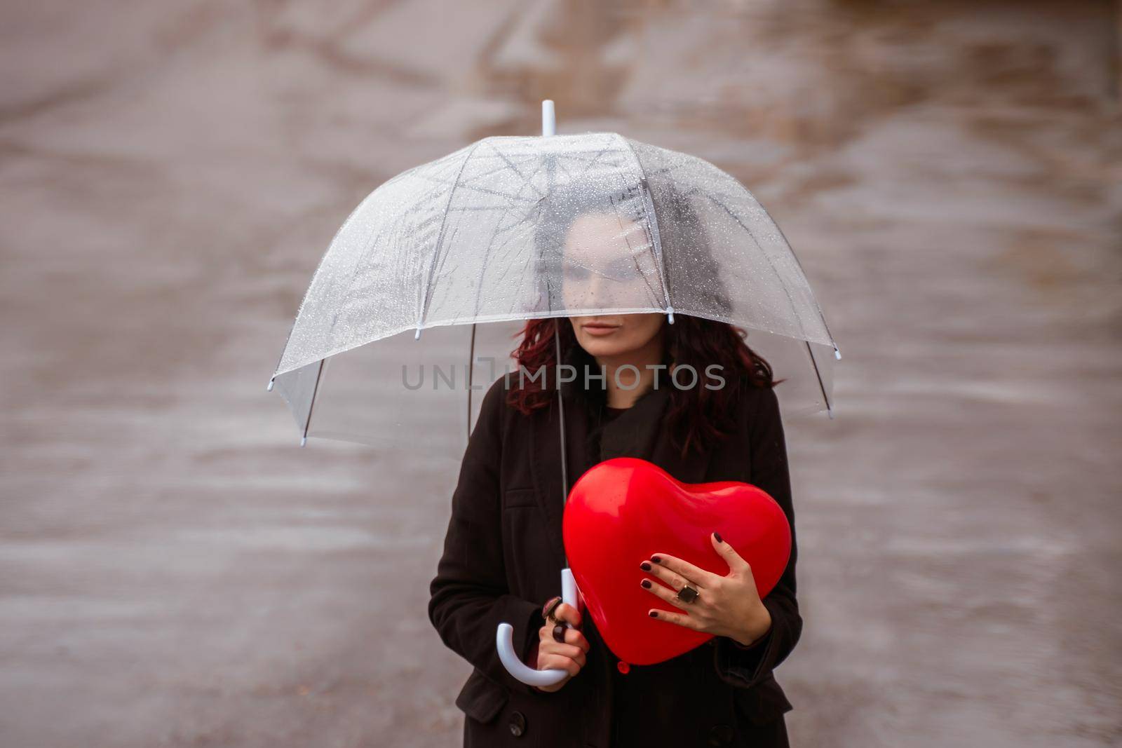 Sad middle-aged woman in a black coat and boots under a transparent umbrella. He holds a balloon in the form of a heart in his hands. Rain in the city, outdoor scene, water drops on the umbrella. The concept of loneliness, unhappy love. by Matiunina