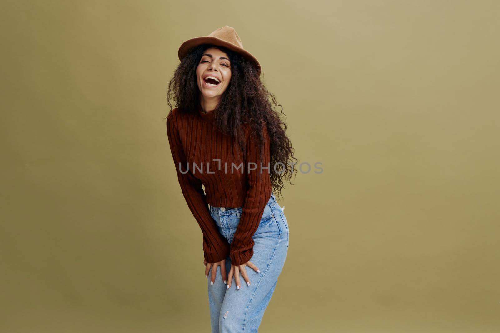 Enjoyed stylish curly Latin female in brown hat, smiling at camera, holding hand on hips, laughing, saying Yes, isolated green background. Copy space clothing fashion brands, free place for your ad.