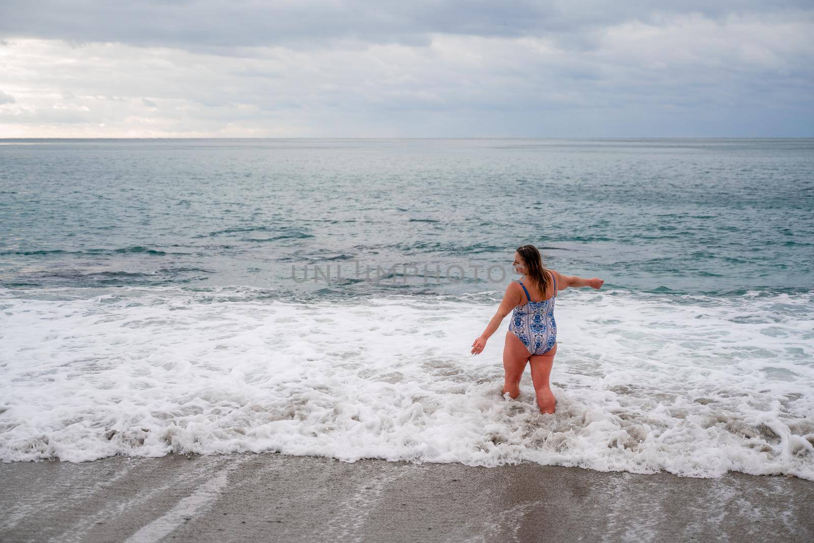 A plump woman in a bathing suit enters the water during the surf. Alone on the beach, Gray sky in the clouds, swimming in winter