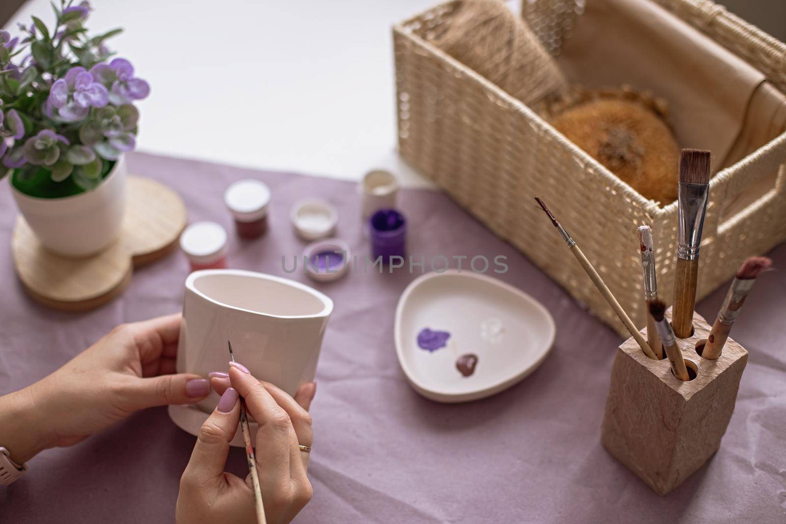 Womens hands are painting a white ceramic flower pot with a brush. On the table are purple craft paper, brushes, paint jars, and a wicker box. Close-up. View from above