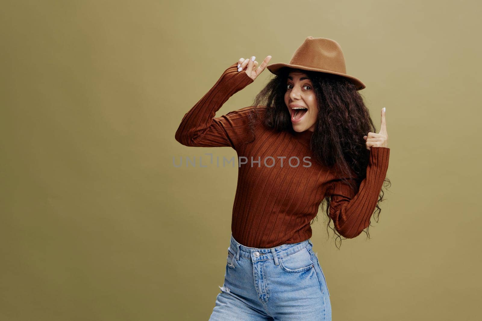 Enjoyed stylish curly Latin female in brown hat, smiling at camera, holding hand on hat, pointing finger up, isolated green background. Copy space clothing fashion brands, free place for your ad.
