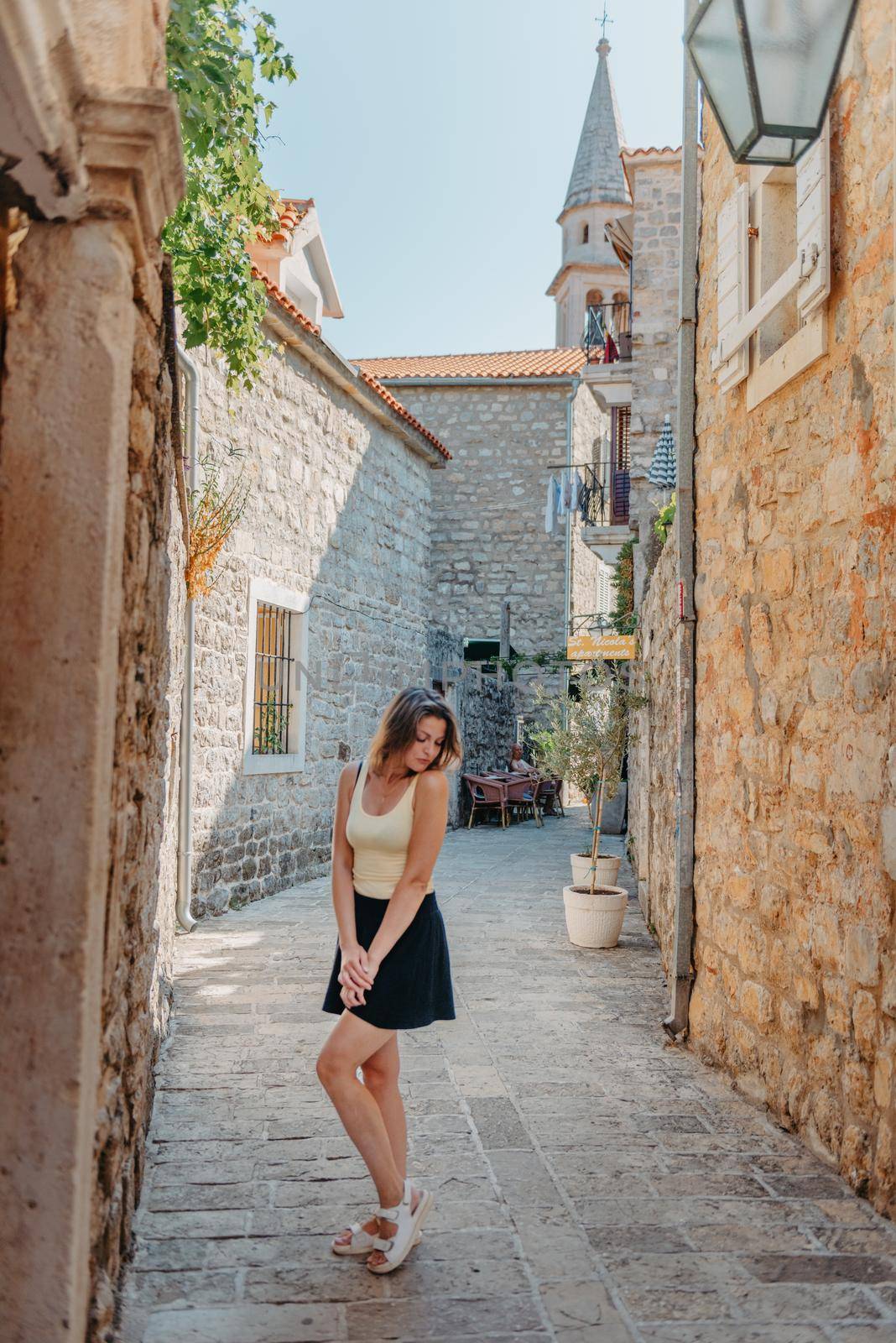 Girl tourist walking through ancient narrow street on a beautiful summer day in MEDITERRANEAN MEDIEVAL CITY , OLD TOWN bUDVA, MONTENEGRO. Young beautiful cheerful woman walking on old street at tropical town. Pretty girl looking at you and smiling