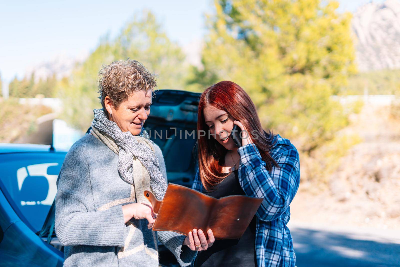 Mother and daughter standing next to the broken down vehicle checking the documents and calling the tow truck. Two friends on holiday have a car accident and have to call the mechanic in the middle of the road. Clear road on a very sunny day next to a forest with a mountain in the background. Red-haired girl calling tow truck and blonde woman looking at insurance papers.