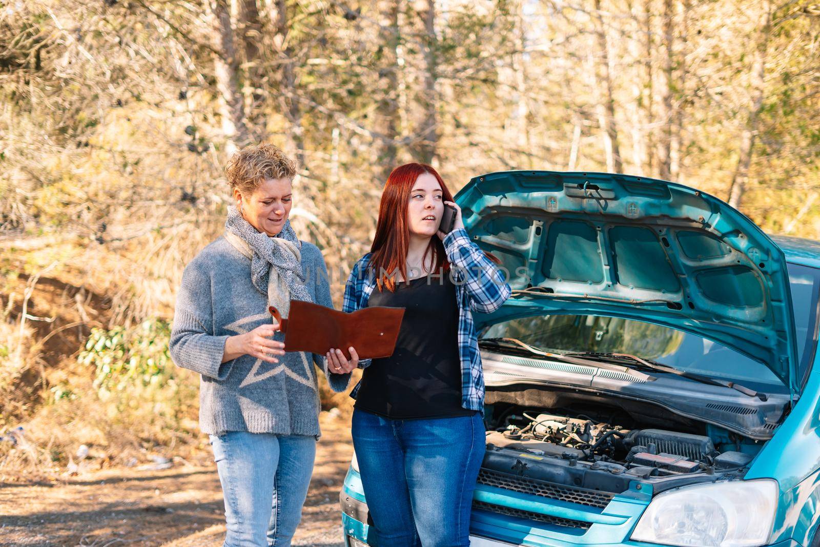 Mother and daughter standing next to the broken down vehicle checking the documents and calling the tow truck. Two friends on holiday have a car accident and have to call the mechanic in the middle of the road. Clear road on a very sunny day next to a forest in the background. Red-haired girl calling tow truck and blonde woman looking at insurance papers.