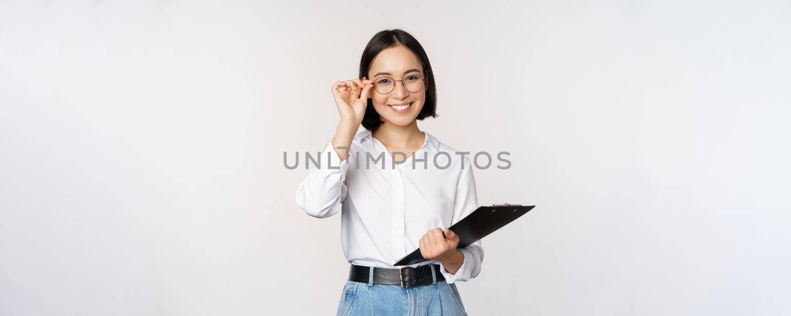 Young woman, office worker manager in glasses, holding clipboard and looking like professional, standing against white background.