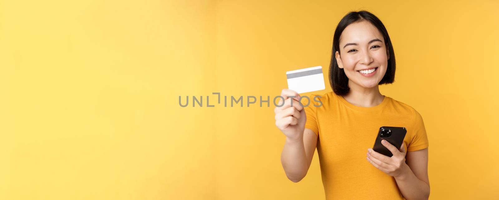 Joyful asian girl smiling, showing credit card and smartphone, recommending mobile phone banking, standing against yellow background.