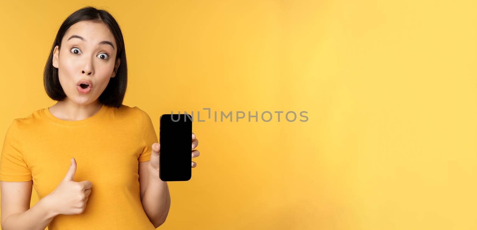 Excited asian woman showing mobile phone screen, thumbs up, like smth good, recommending smartphone application, standing over yellow background.