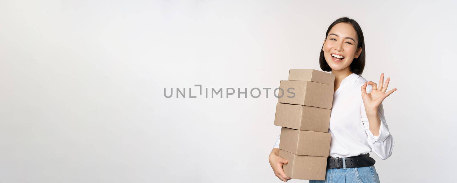 Portrait of beautiful happy woman carry boxes with orders. Businesswoman with carry delivery and showing okay sign, white background by Benzoix