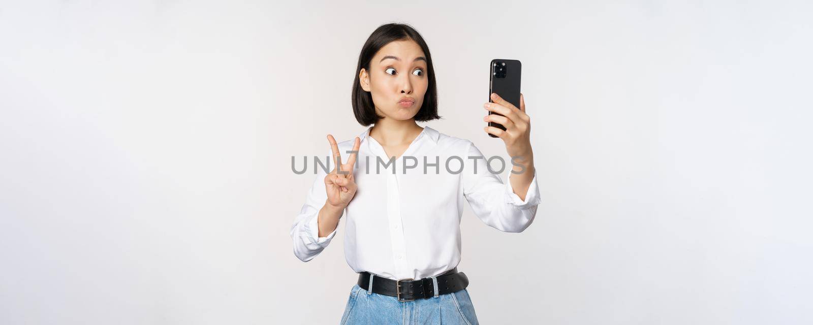 Funny asian girl showing peace, v-sign at smartphone camera, posing for photo, taking pictures with mobile application, standing over white background.