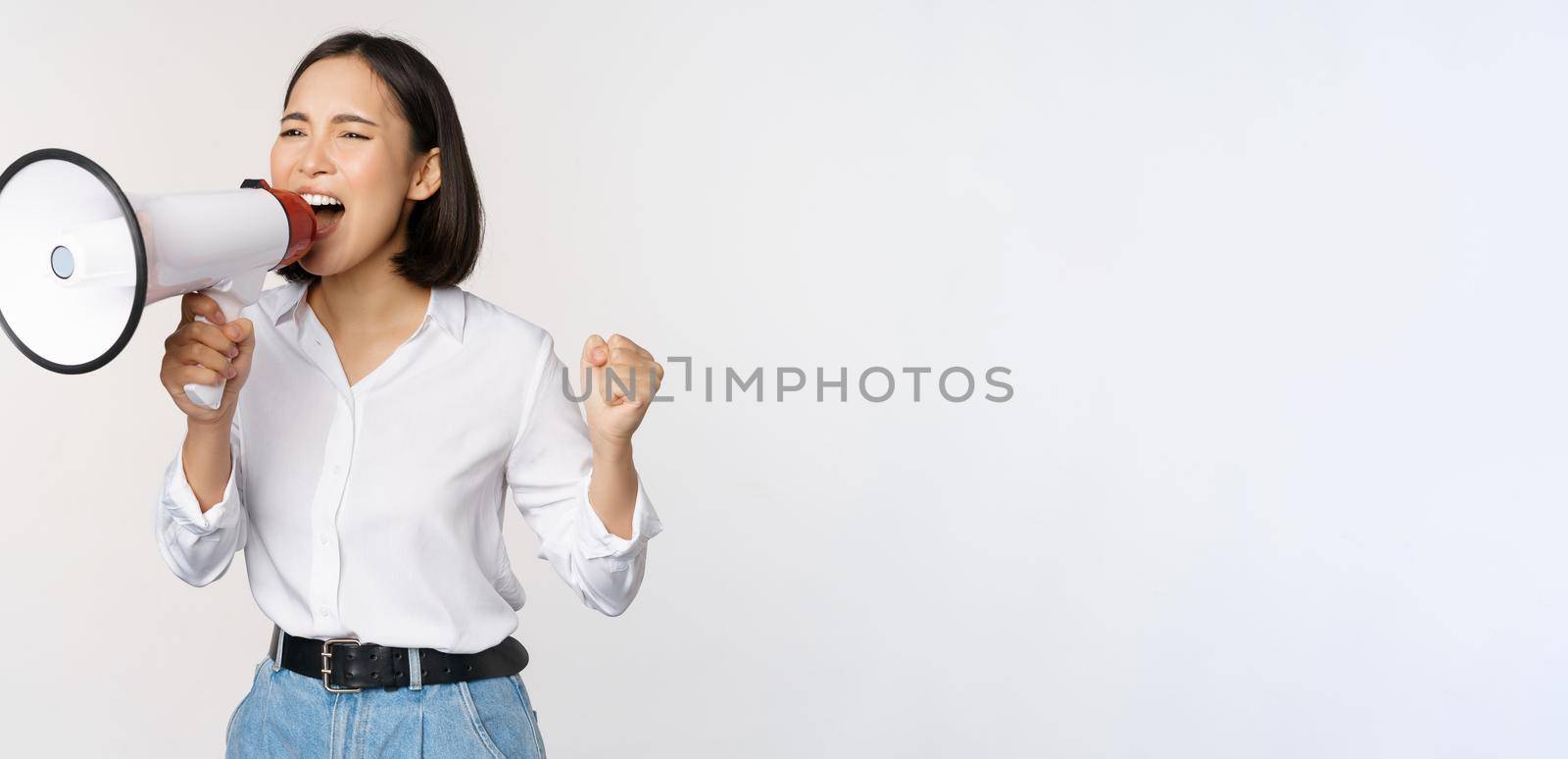 Enthusiastic asian woman, girl activist shouting at protest, using megaphone, looking confident, talking in loudspeaker, protesting, standing over white background by Benzoix