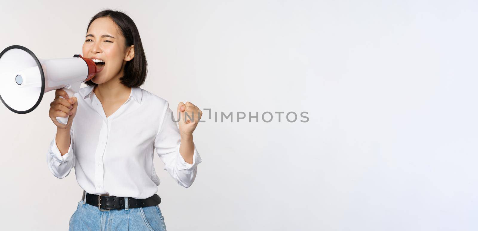 Enthusiastic asian woman, girl activist shouting at protest, using megaphone, looking confident, talking in loudspeaker, protesting, standing over white background.