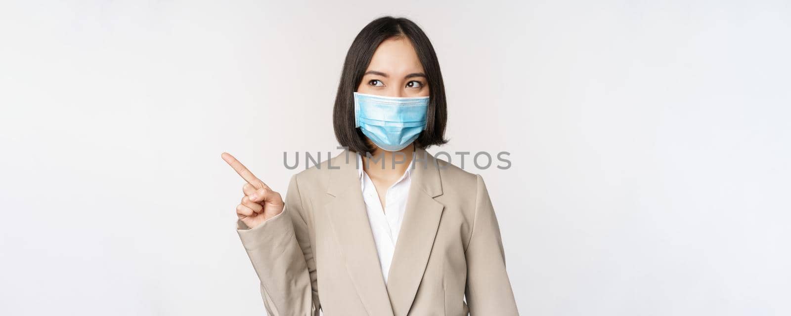 Enthusiastic businesswoman pointing fingers left, wearing medical face mask from covid-19 pandemic, standing over white background.