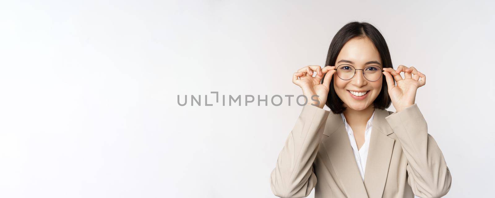 Smiling asian businesswoman trying new glasses, wearing eyewear, standing in suit over white background by Benzoix