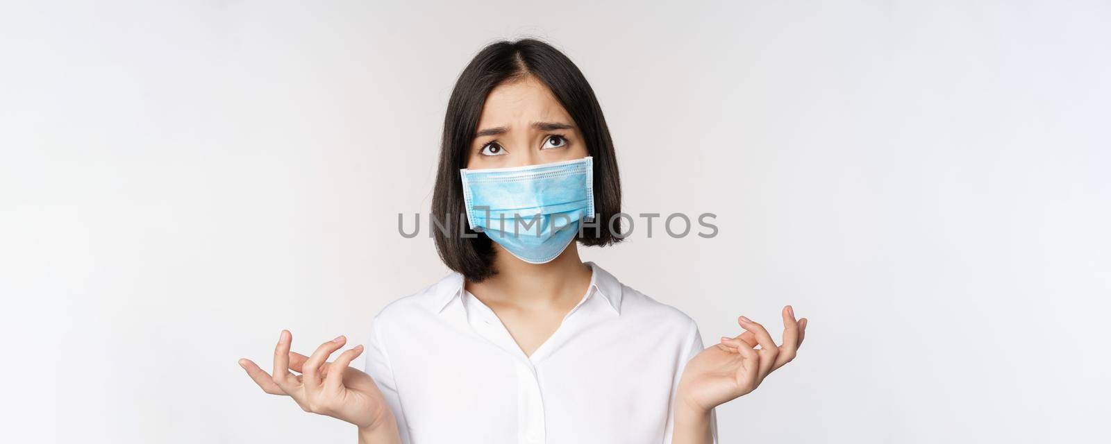 Distressed and miserable young asian woman in face mask, looking up, looking up sad, standing over white background.