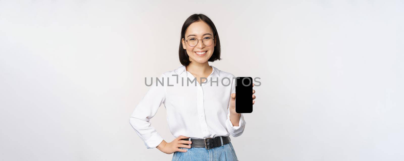 Smiling korean woman showing smartphone screen, demonstrating mobile application, standing over white background.