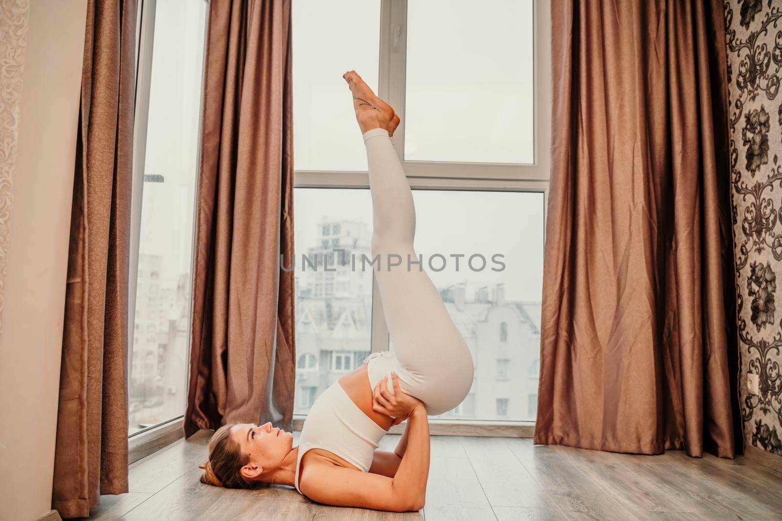 Young athletic attractive woman practicing yoga. Works out at home or in a yoga studio, sportswear, white pants and a full-length top indoors. Healthy lifestyle concept.
