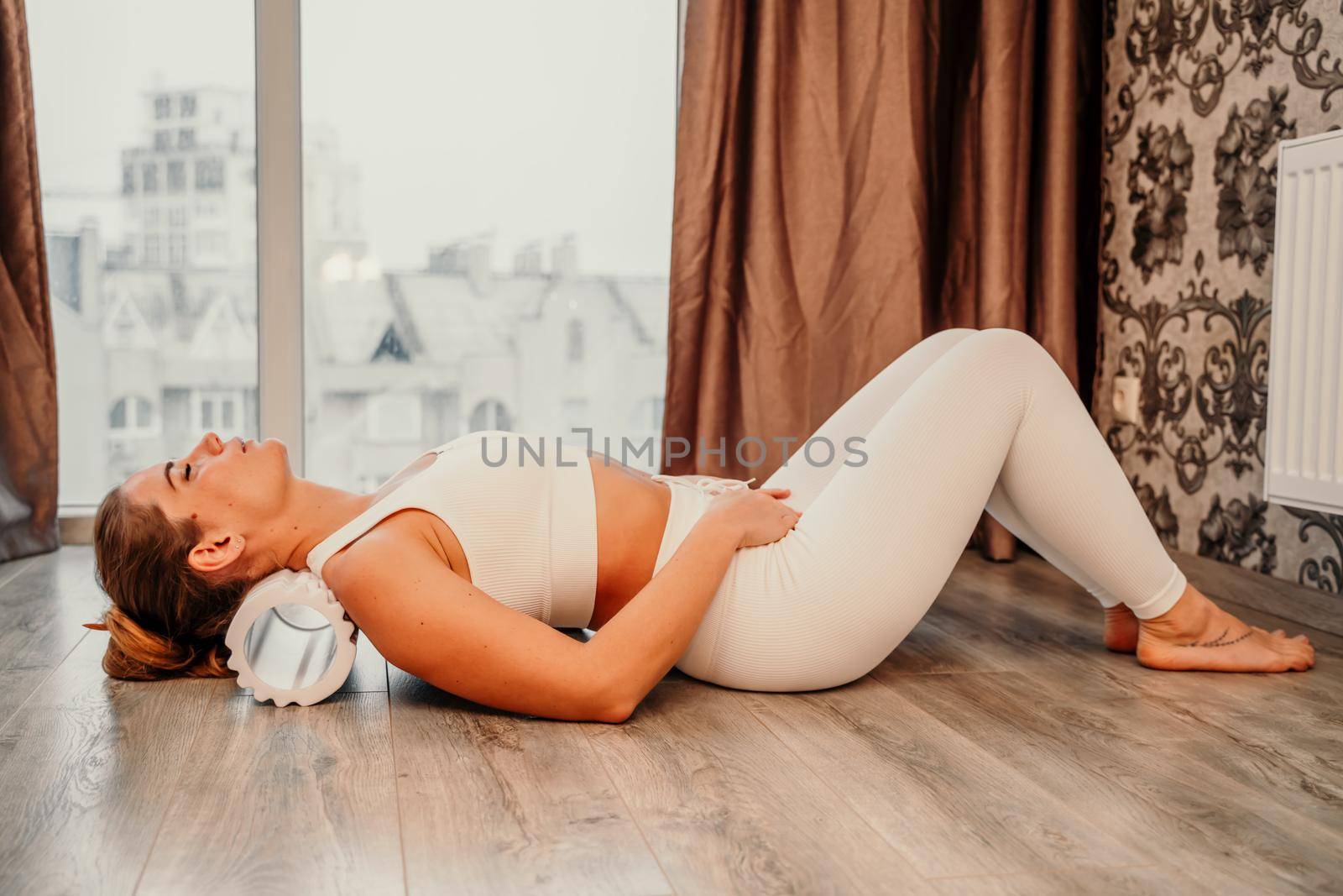 Adult athletic woman, in white bodysuit, performing fascia exercises on the floor - caucasian woman using a massage foam roller - a tool to relieve tension in the back and relieve muscle pain - the concept of physiotherapy and stretching training by Matiunina