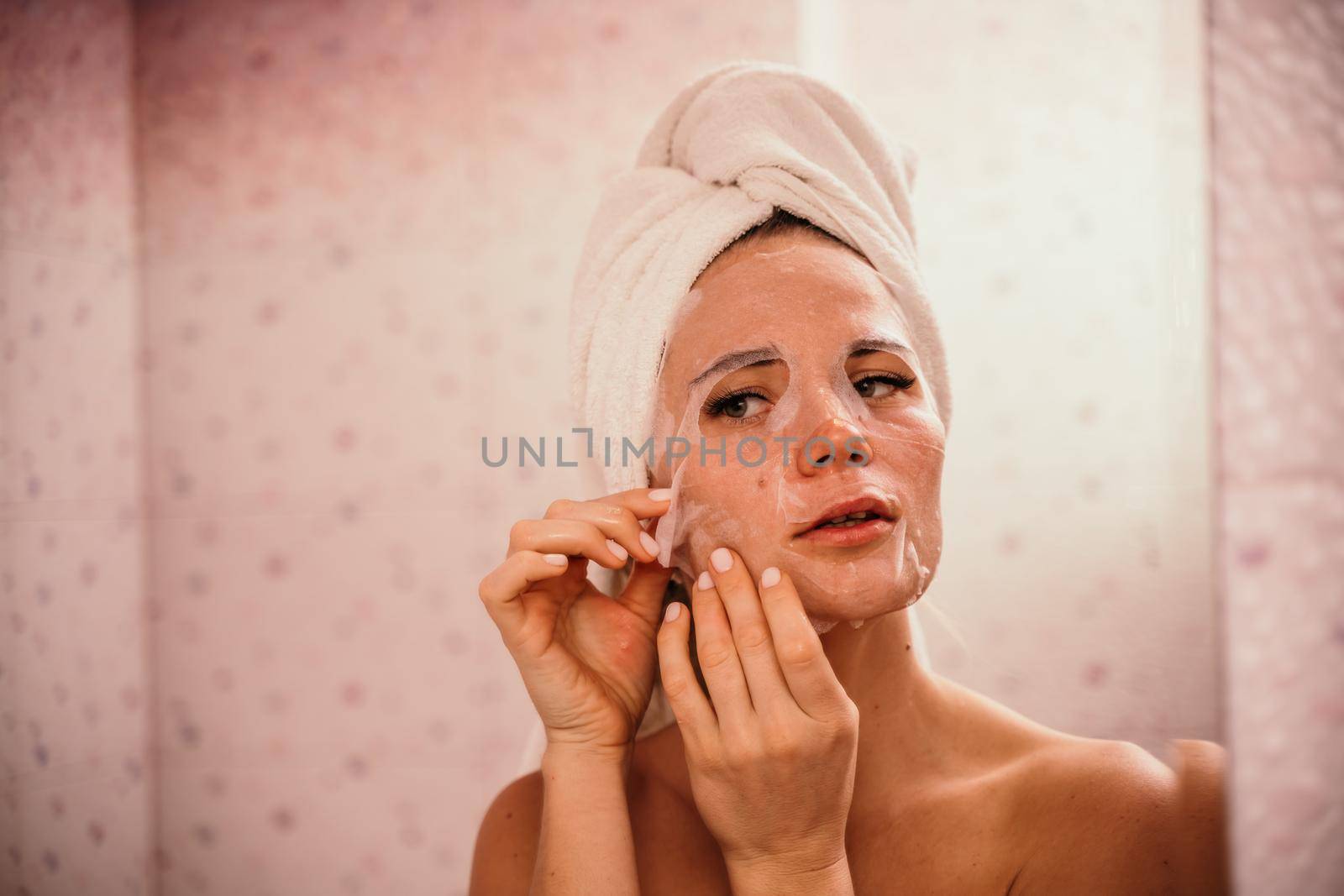 Young beautiful woman using a moisturizing facial mask after taking a bath. Pretty attractive girl in a towel on her head stands in front of a mirror in a home bathroom. Daily hygiene and skin care.