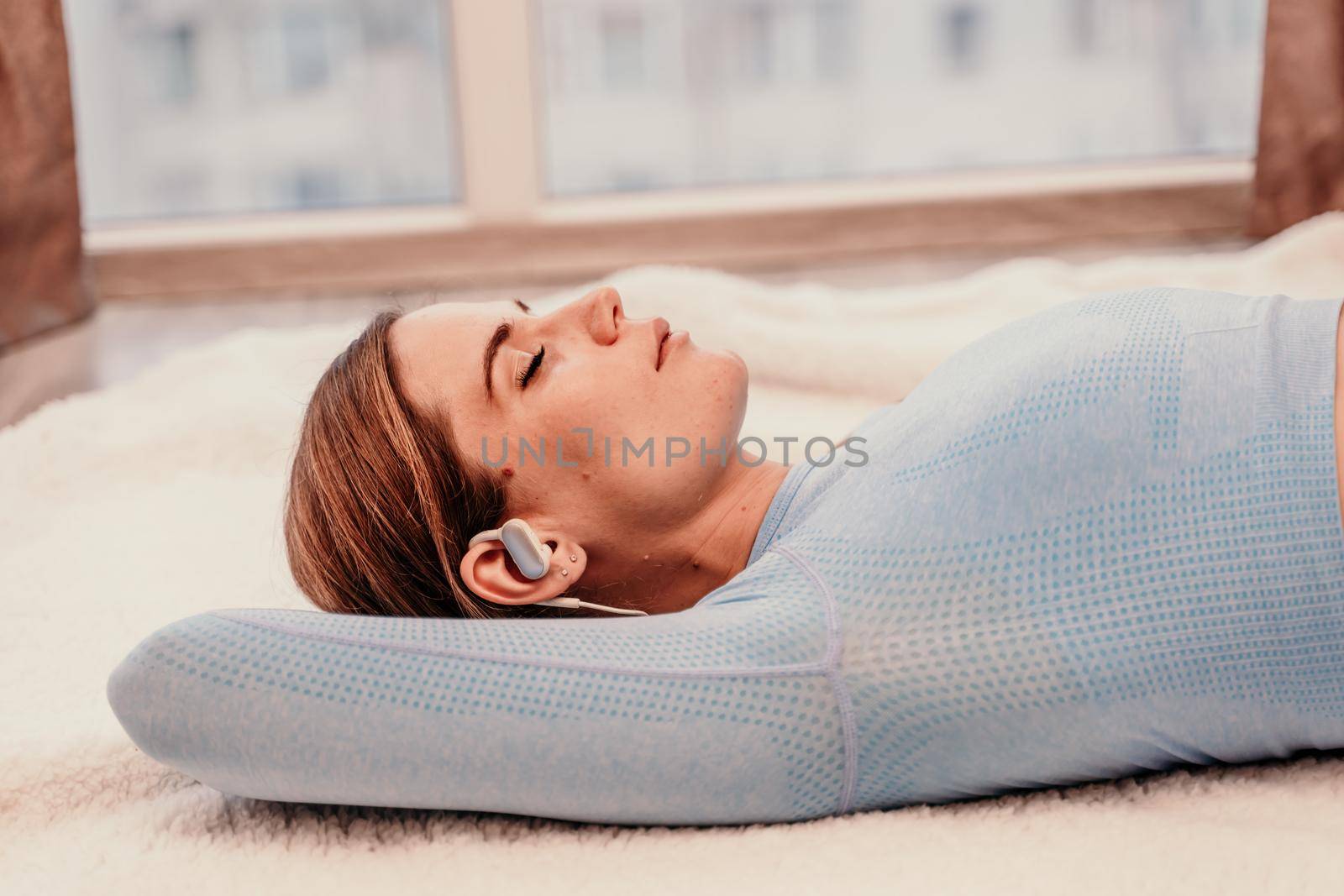 Side view portrait of relaxed woman listening to music with headphones lying on carpet at home. She is dressed in a blue tracksuit