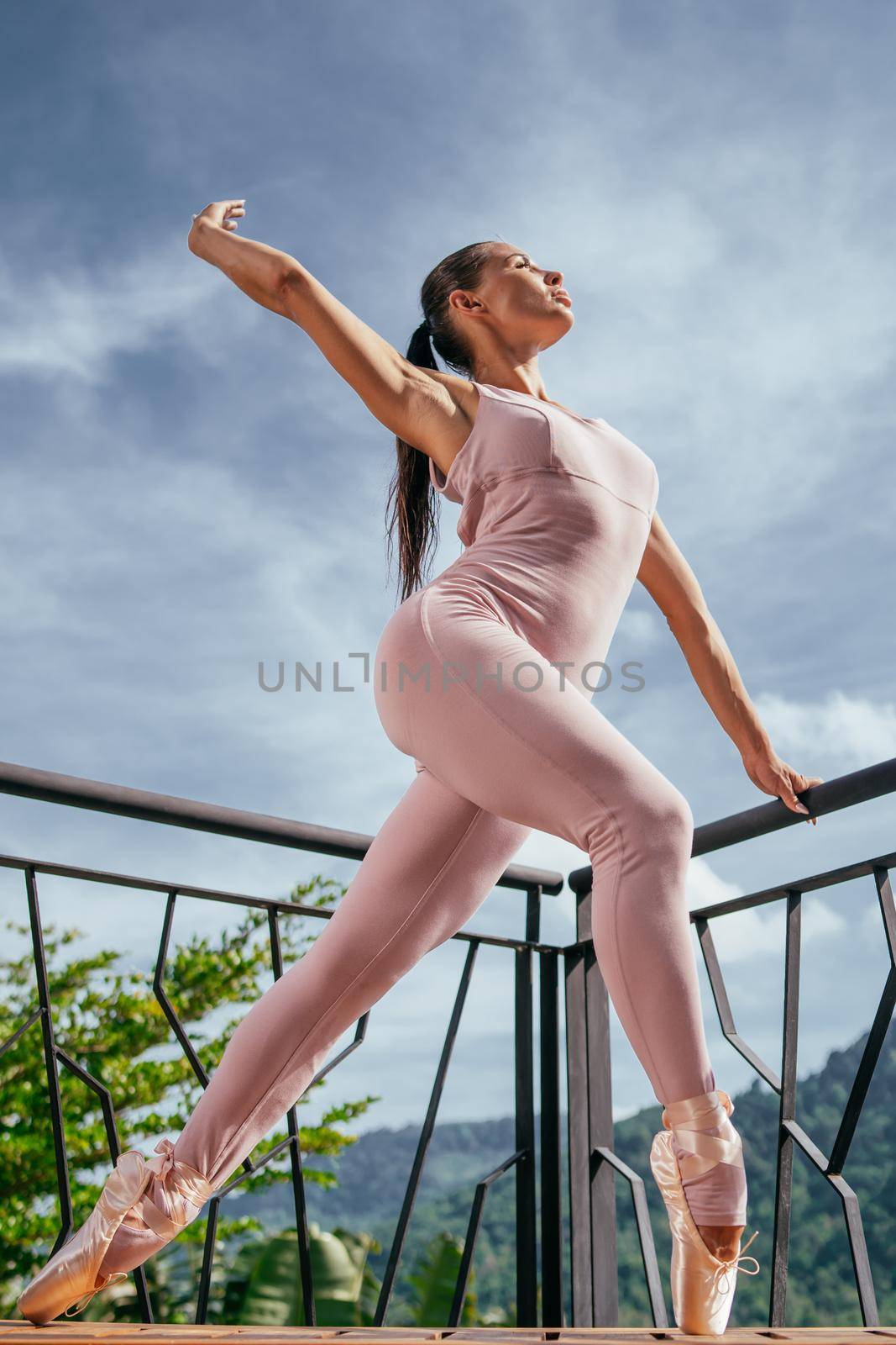 Young fit ballerina in pink pointes stretching and standing on toes in warm sunrise against blue sky. Beautiful ballerina dancing in ballet shoes in outdoor. Beauty fasion. Power, wellbeing concept.