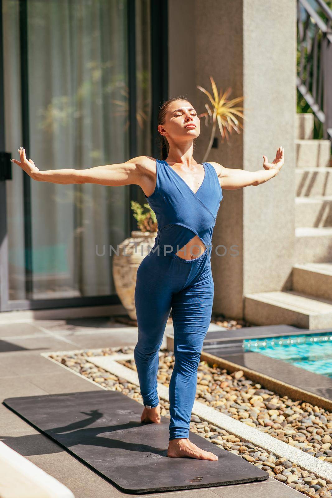 Fit sporty young woman in blue jumpsuit is stretching, warming up after workout by XGroup