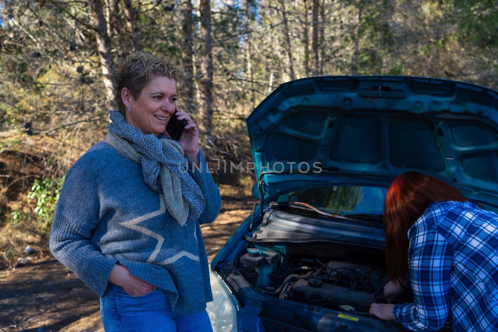 blonde adult woman and red-haired young woman calling the emergency service on the road in front of a forest with the bonnet open due to car breakdown, dressed in blue jumper and blue chequered shirt.