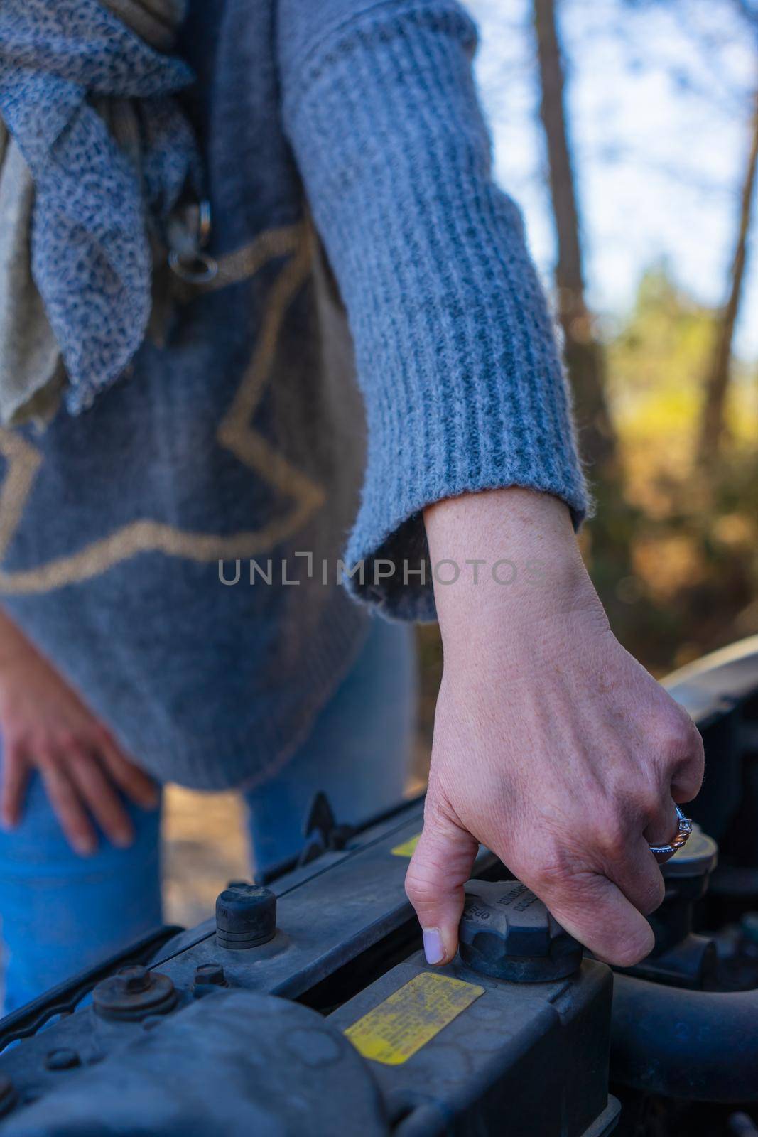 close-up of the hand of an adult woman repairing a car engine, with the bonnet open, wearing a blue jumper and blue jeans.