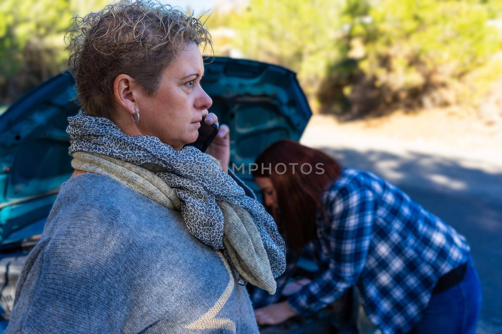 blonde adult woman and red-haired young woman calling the emergency service on the road in front of a forest with the bonnet open due to car breakdown, dressed in blue jumper and blue chequered shirt.