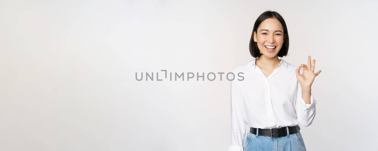 Very good. Smiling asian woman showing okay with satisfied face expression, praise and compliment great job, pleased by smth, standing over white background.