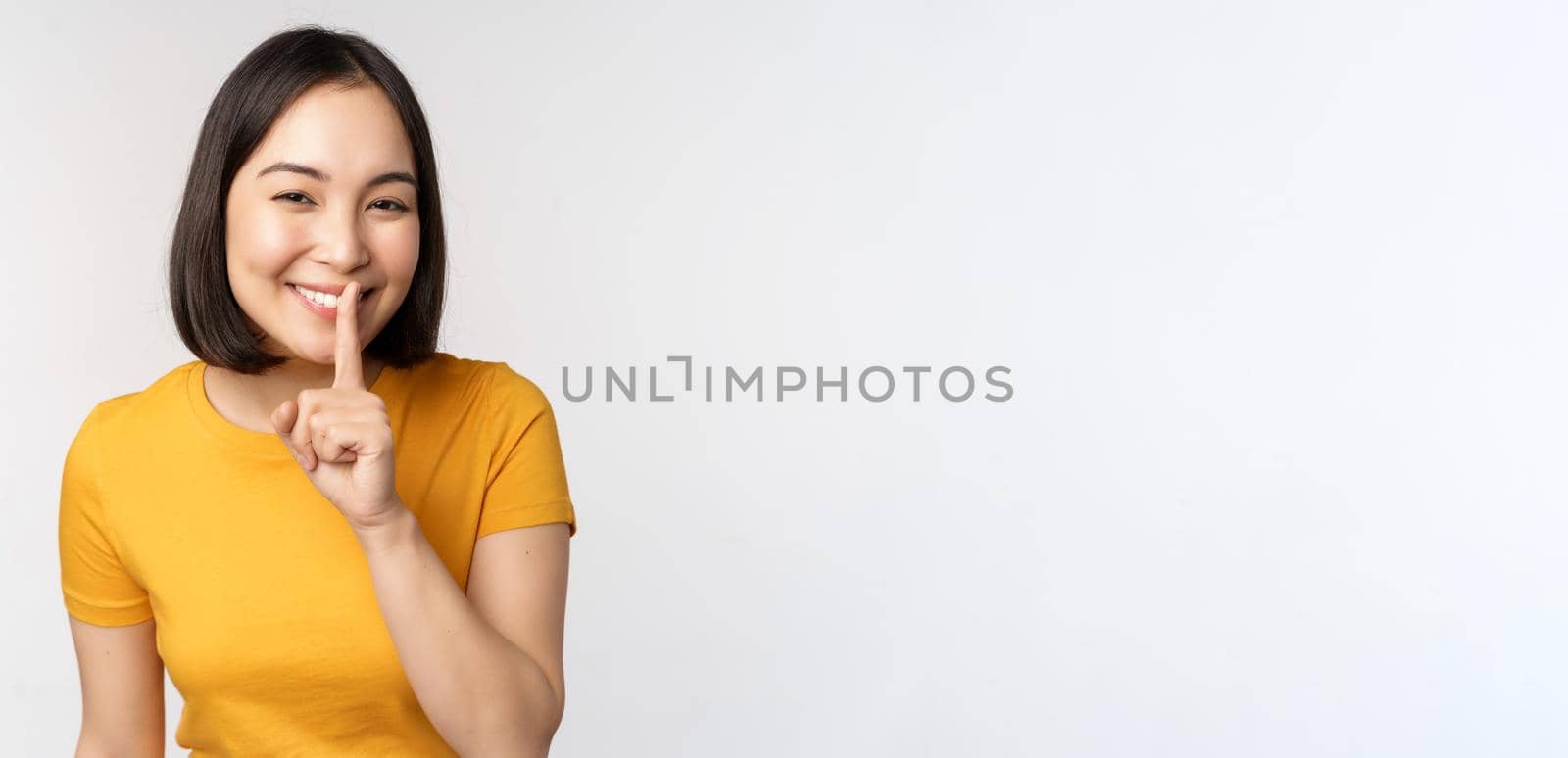Keep quiet. Cute asian woman make shhh gest, showing shush, hush sign, press finger to lips, silence, standing over white background by Benzoix