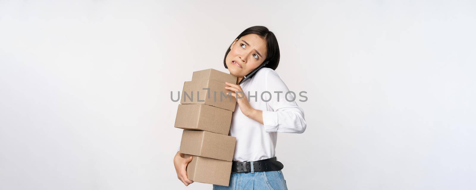 Young asian businesswoman answer phone call, talking on mobile while carrying pile of boxes with orders, standing over white background.