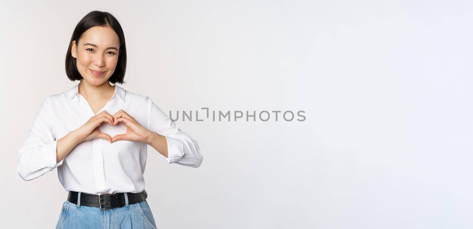 Love gesture. Beautiful asian young woman, showing heart gesture and smiling, express care and affection, standing over white background.