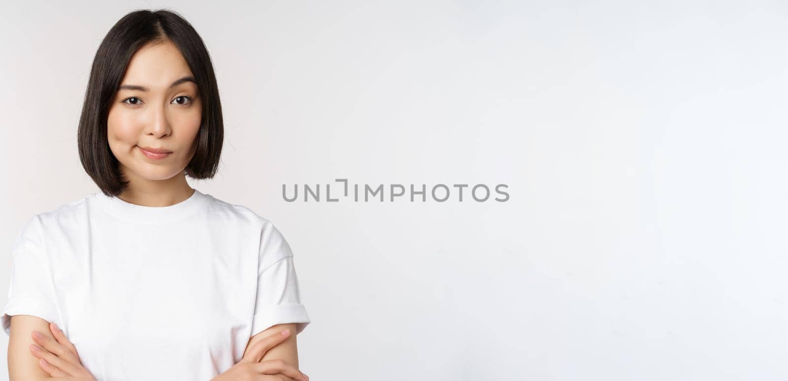 Close up of korean girl, looking skeptical, cross arms on chest and smirk, stare with disbelief at camera, standing over white background.