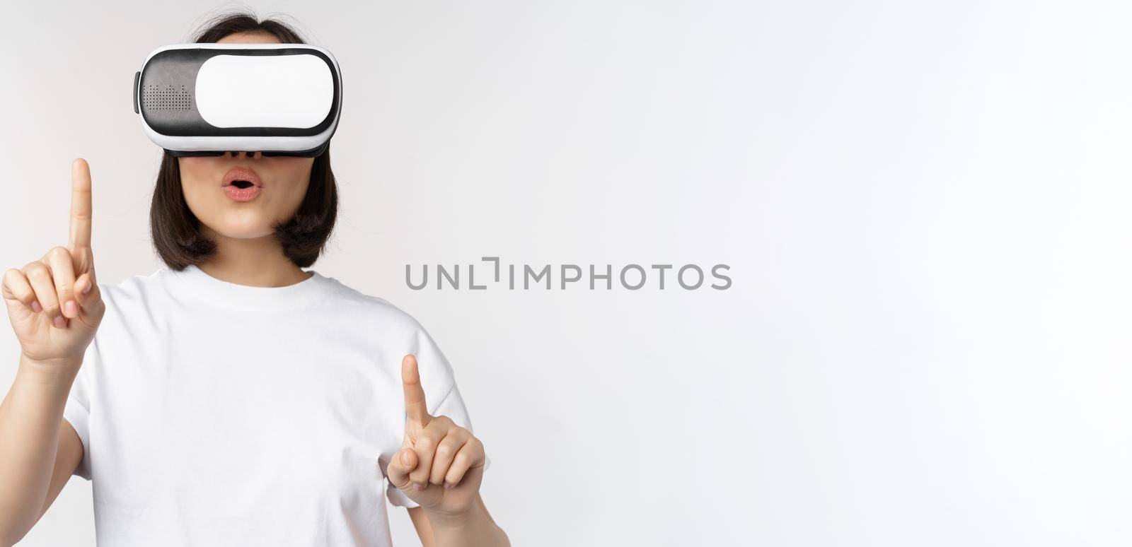 Potrait of asian woman in virtual reality glasses, pointing, choosing smth in VR headset, standing over white background.