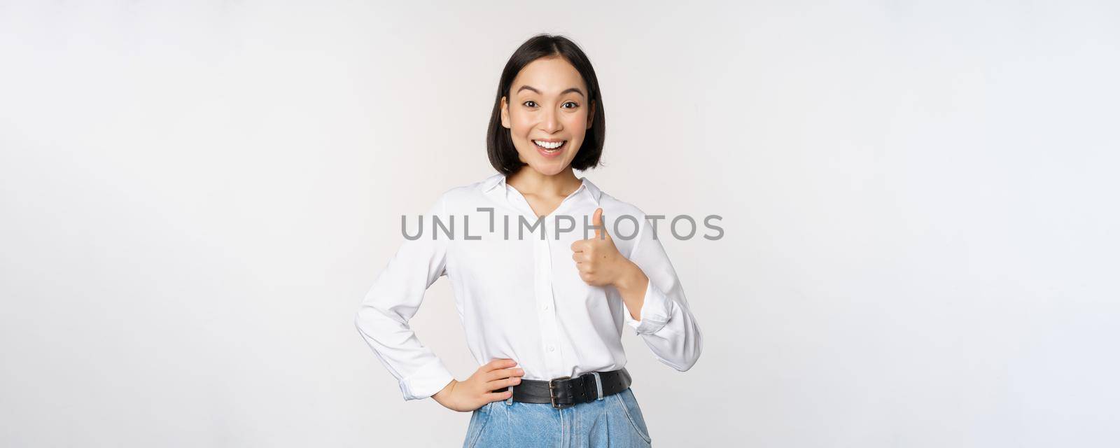 Very good. Smiling enthusiastic asian female entrepreneur, showing thumb up in approval, praise excellent job, satisfied with results, standing over white background.