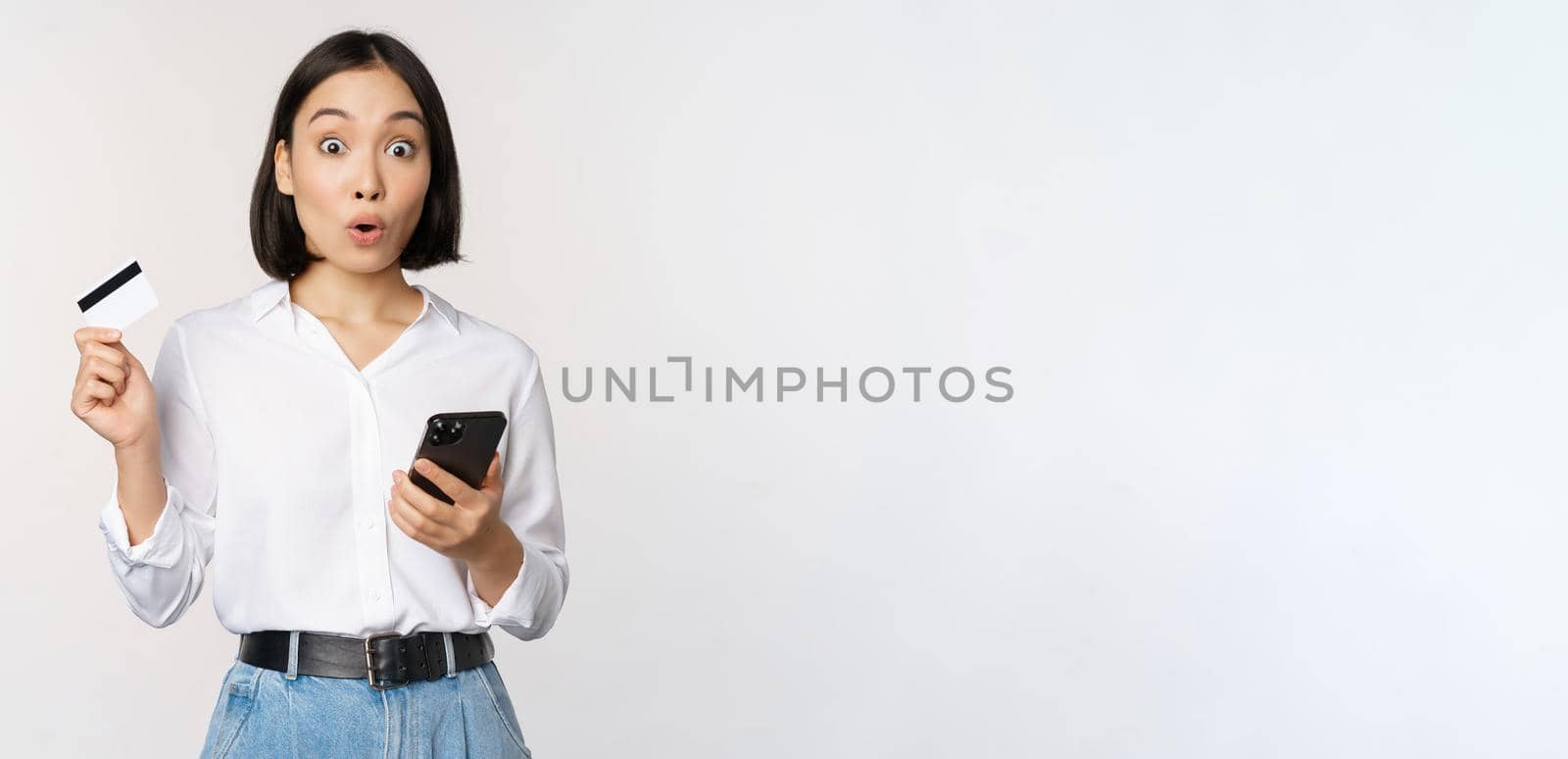 Online shopping concept. Image of surprised asian girl, holding credit card and smartphone, looking amazed in disbelief at camera, white background by Benzoix