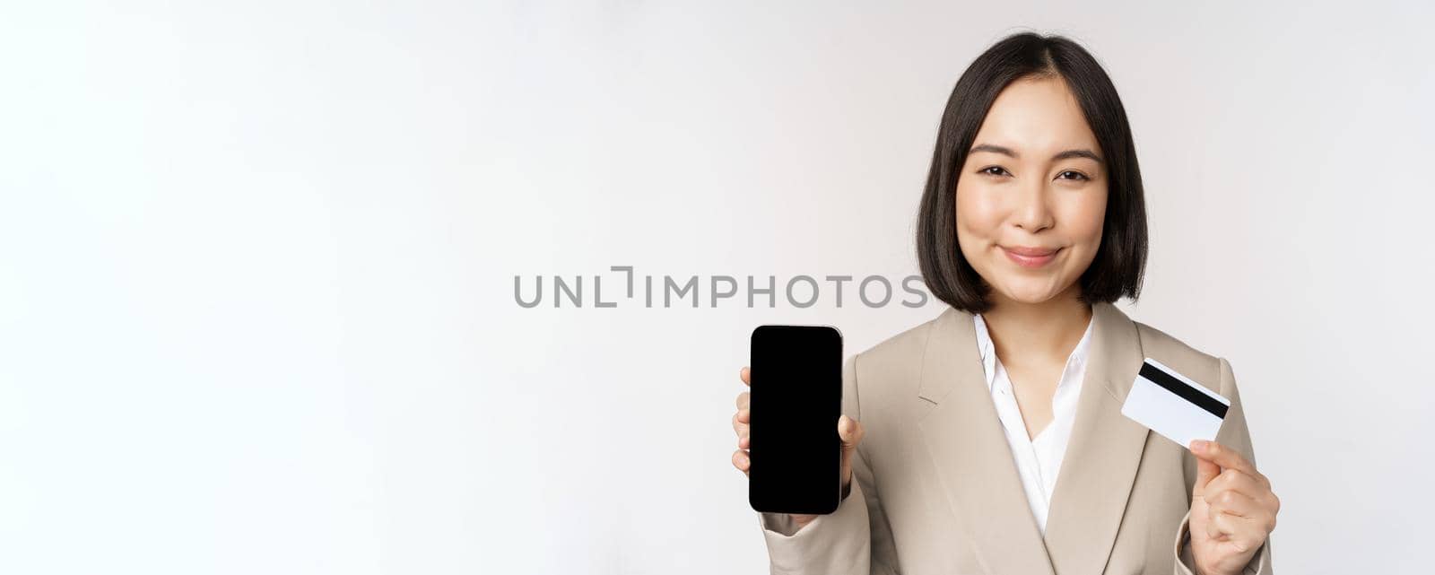 Smiling corporate woman in suit, showing mobile phone screen and app on mobile phone, smartphone screen, standing over white background.