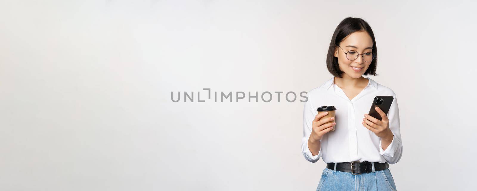 Image of modern asian woman looking at mobile phone, drinking takeaway coffee, wearing glasses, using smartphone app, standing over white background.