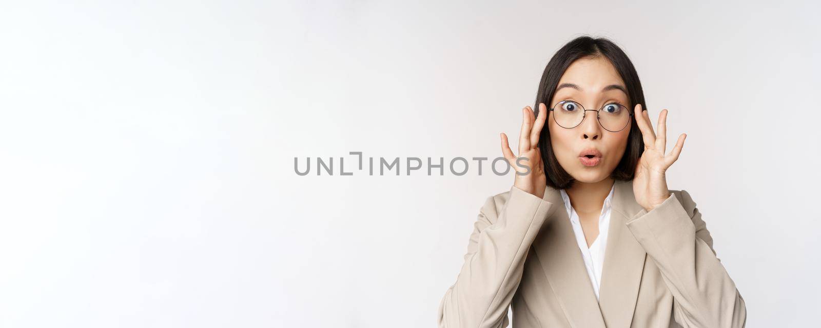 Portrait of asian businesswoman in glasses, looking surprised amazed at camera, standing in suit over white background.